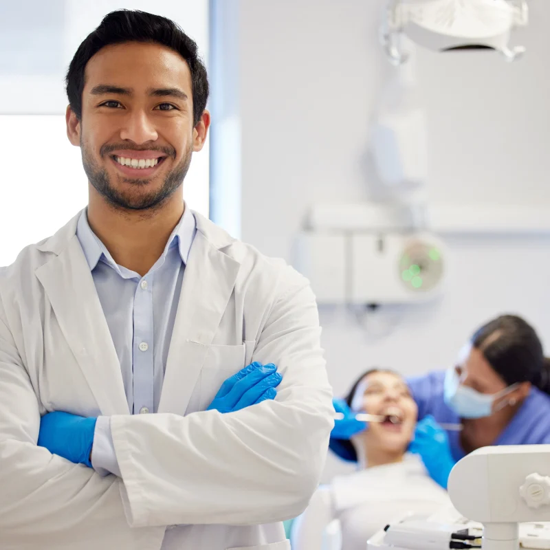 Smiling dentist in clinic with patient in background receiving orthodontic treatment